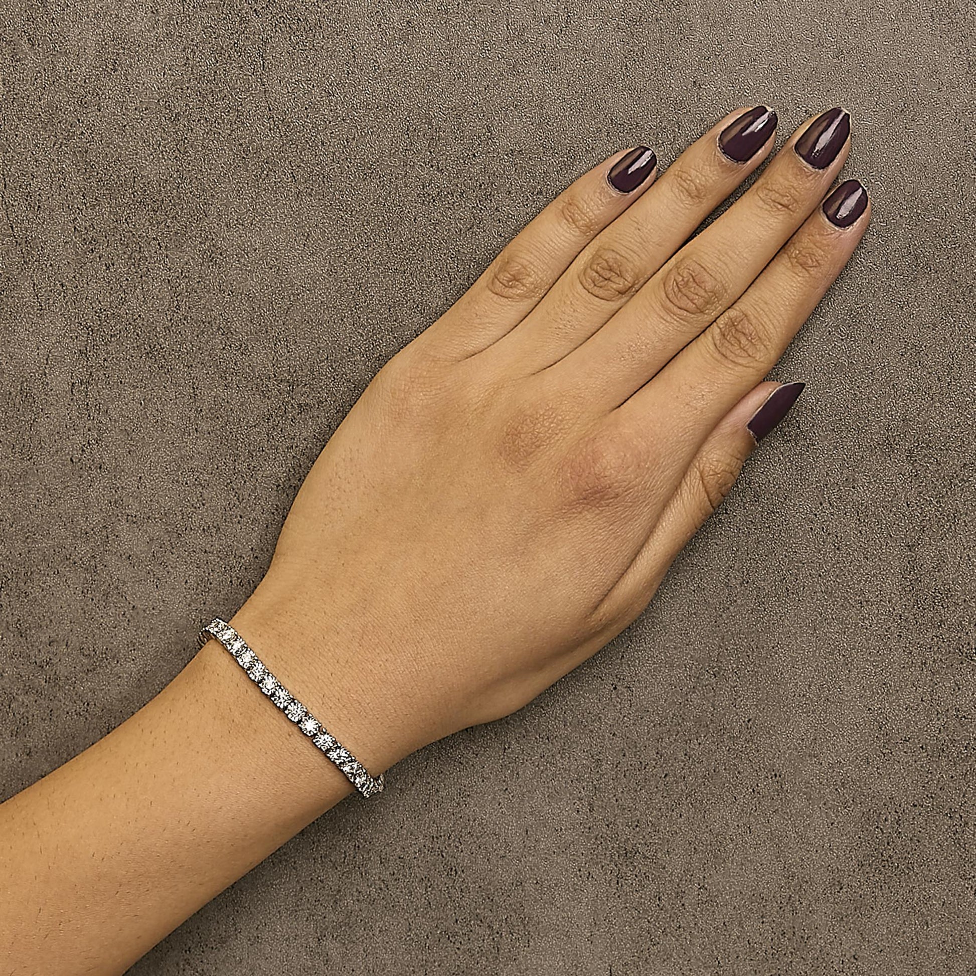 Top-down view of a white metal bracelet with round white gemstones in miracle setting, worn on a model's wrist against a textured surface.