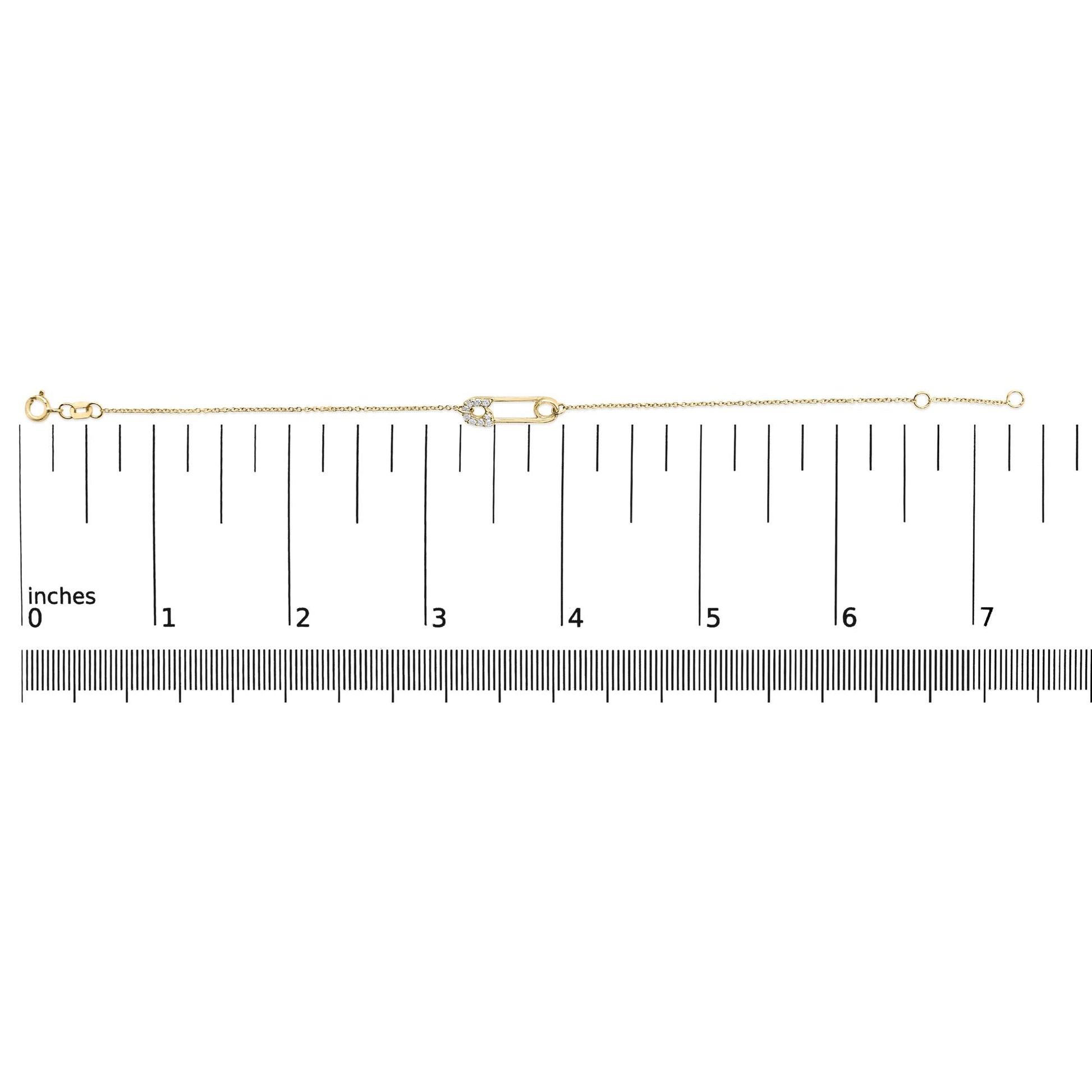 Yellow metal bracelet with a safety pin charm adorned with white round gemstones in prong settings, shown on a measuring scale from above.