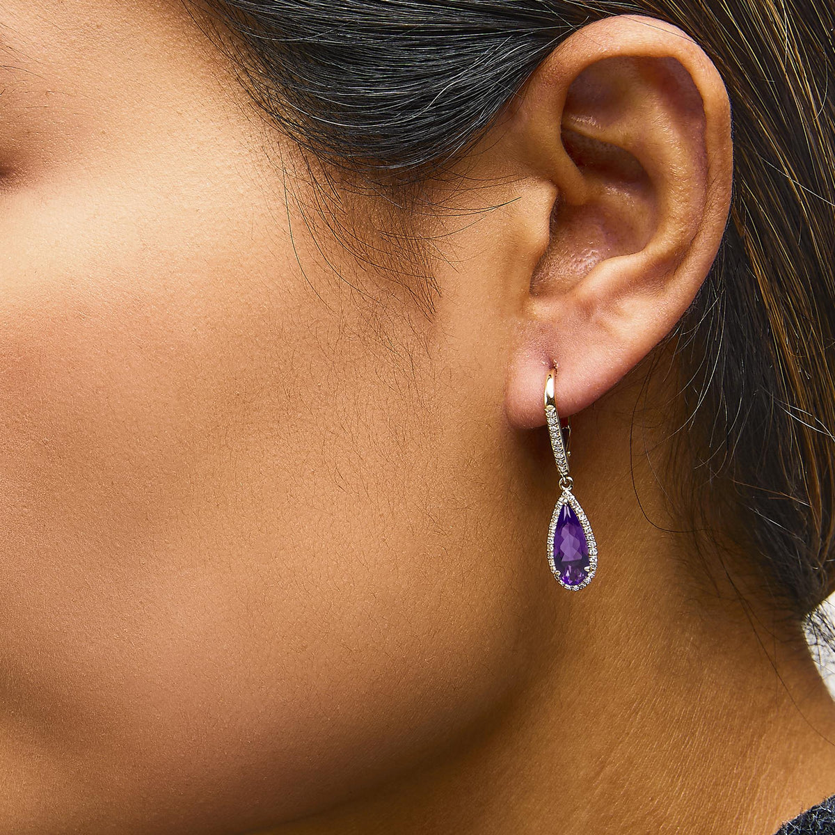 Close-up side view of a model wearing yellow metal drop earrings featuring pear-shaped purple gemstones surrounded by small white gemstones.