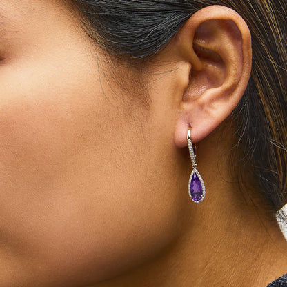 Close-up side view of a model wearing yellow metal drop earrings featuring pear-shaped purple gemstones surrounded by small white gemstones.