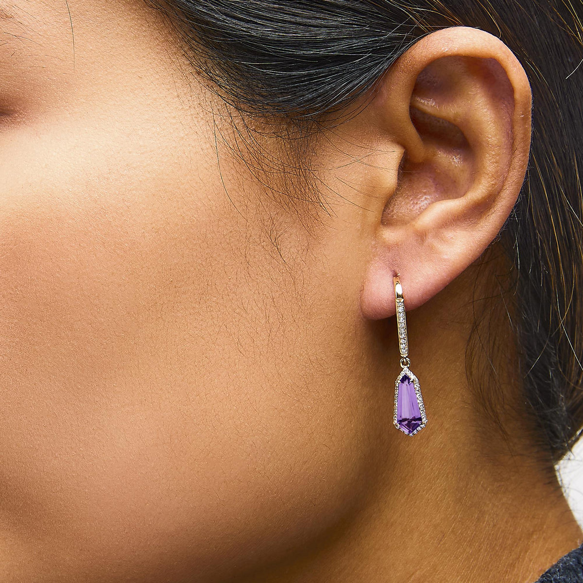 Yellow metal drop earring with round white gemstones in micro-pave setting along the hoop and a dangling purple gemstone, shown worn on a model in close-up side view.