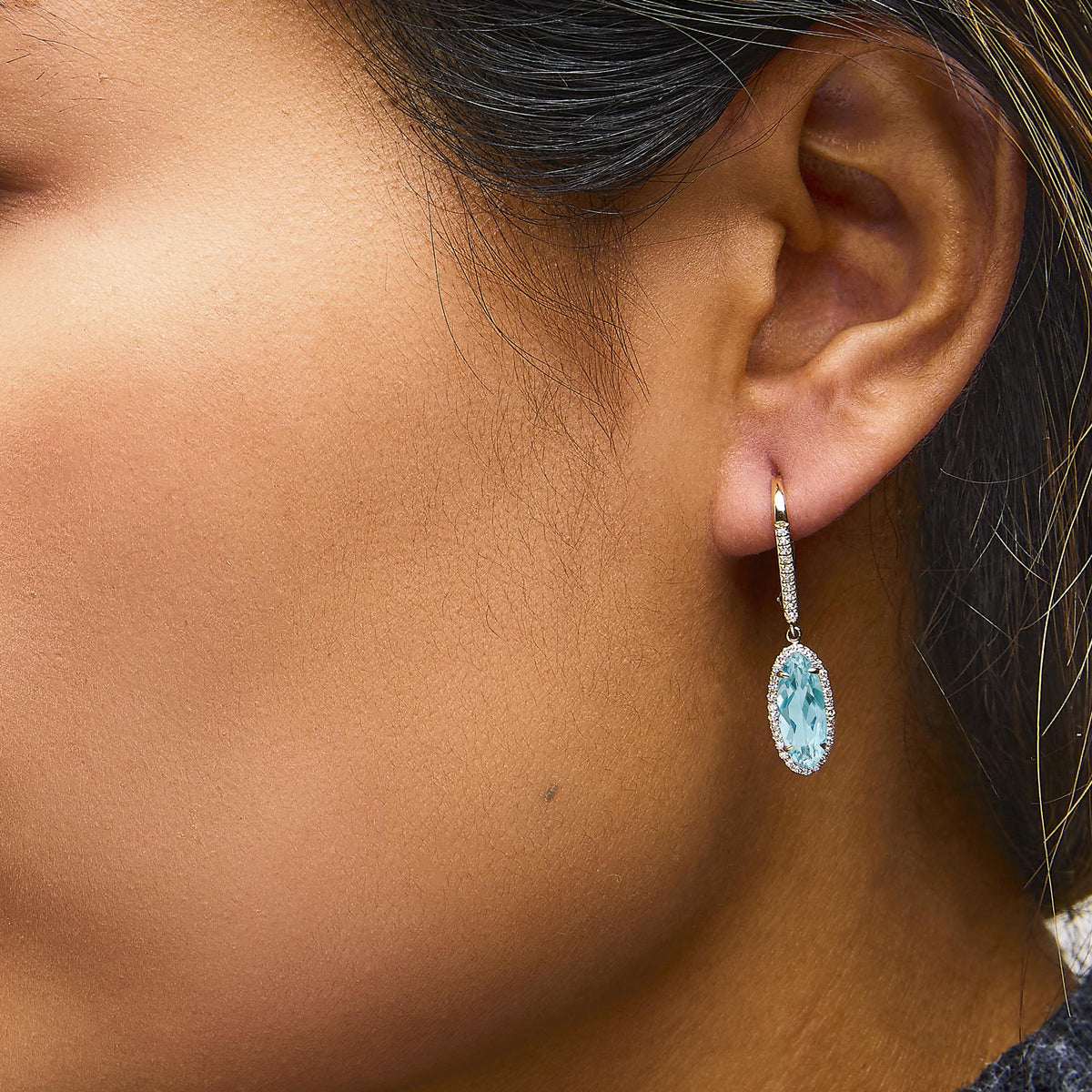 Close-up side angle of a model wearing a yellow metal drop earring featuring a large oval blue gemstone surrounded by small white gemstones in a micro-pave setting.