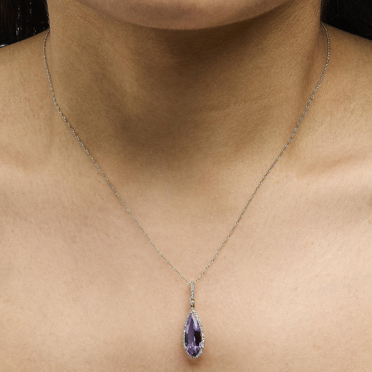 Yellow metal necklace with a pear-shaped purple gemstone surrounded by small white gemstones in a micro-pave setting, shown worn on a model in a close-up front view.