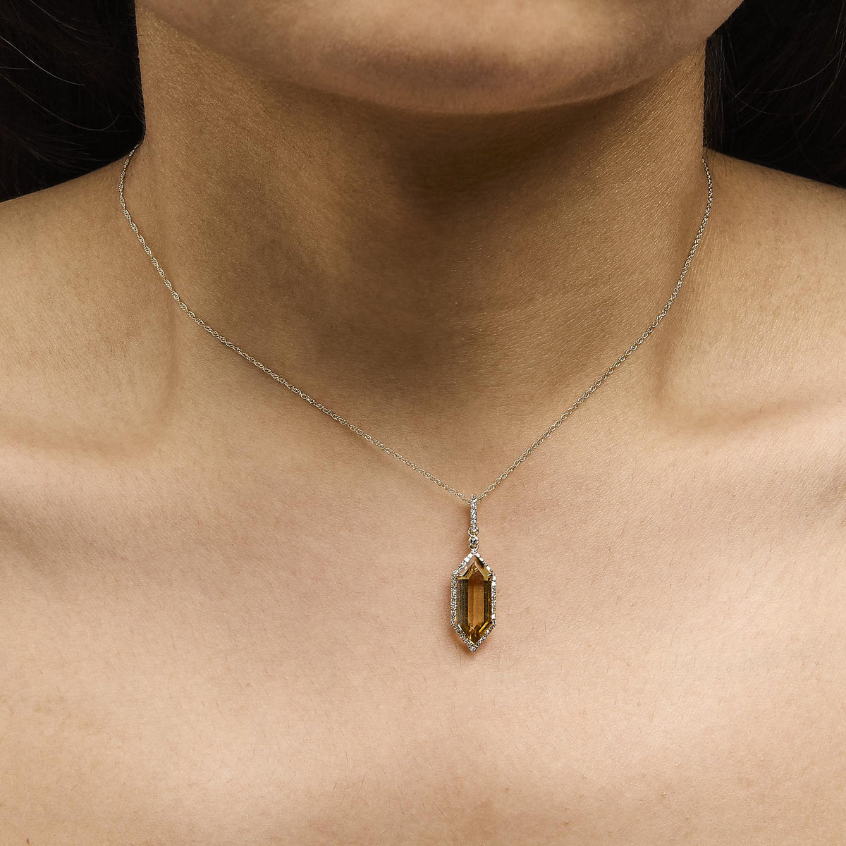 Yellow metal necklace with a vertically elongated hexagonal yellow gemstone pendant, surrounded by small white gemstones, shown worn on a model in a close-up front angle shot.
