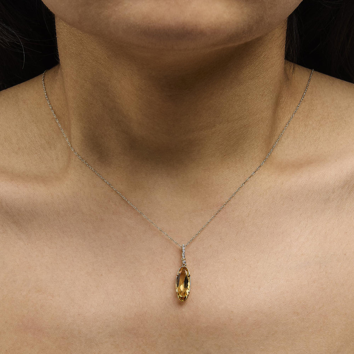 Close-up front view of a model wearing a yellow metal necklace with an oval yellow gemstone pendant accented by small round white gemstones in a vertical arrangement above it.