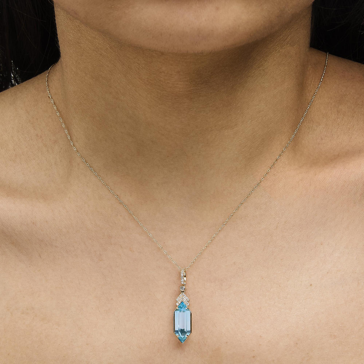 Yellow metal necklace with a rectangular blue gemstone pendant surrounded by small white gemstones in a micro-pave setting, shown worn on a model, close-up angle.