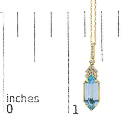 Close-up image of a yellow metal necklace pendant shown on a scale, featuring a vertical row of small white round gemstones in a micro-pave setting, a single blue gemstone in a prong setting, and a large blue gemstone at the bottom.