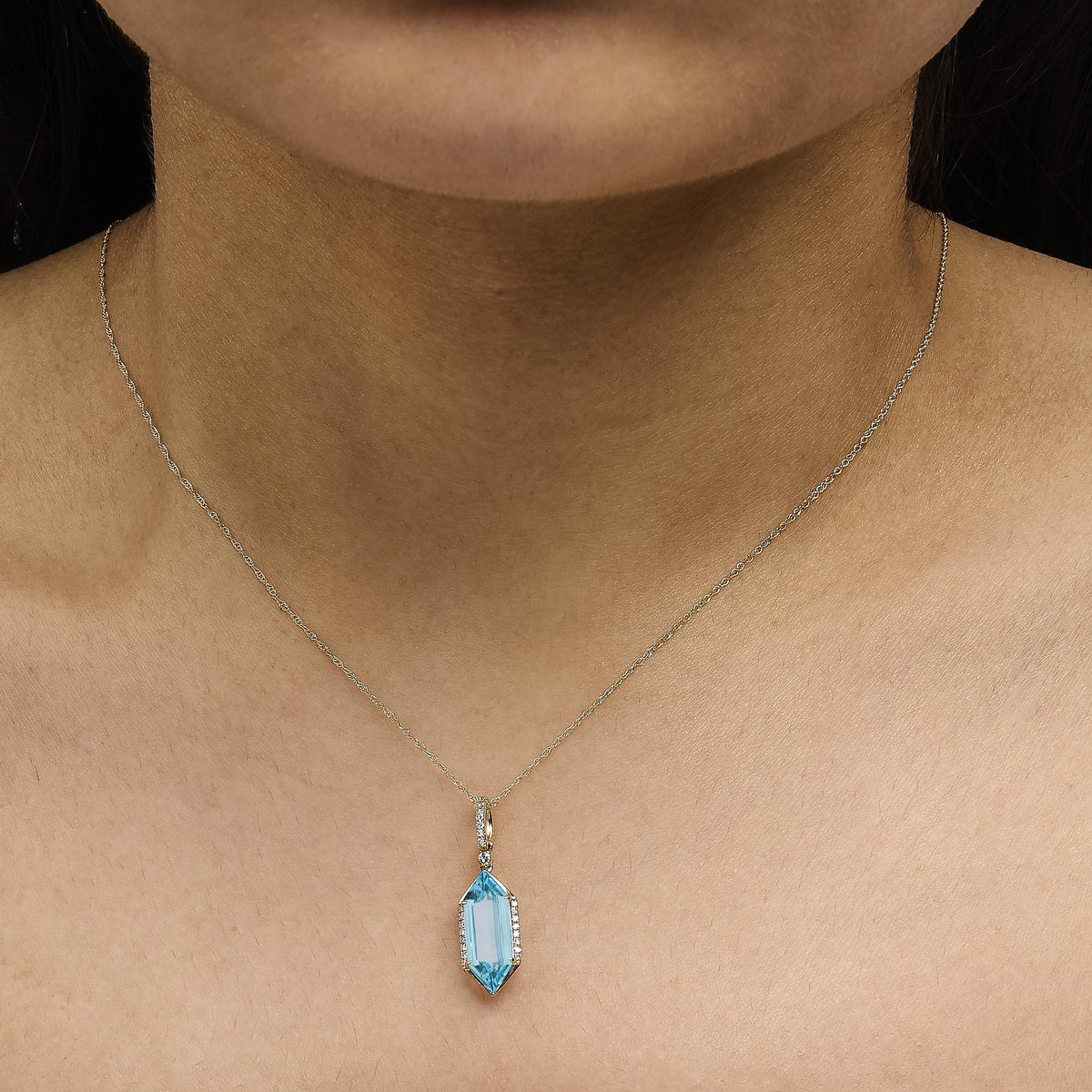 Close-up front view of a model wearing a yellow metal necklace with a pendant featuring a large blue gemstone in a prong setting surrounded by small white gemstones in a micro-pave setting.