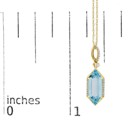 Close-up image of a yellow metal necklace pendant shown on a scale, featuring a geometric blue gemstone with a prong setting, surrounded by small white gemstones in a micro-pave setting along the edges and on a circular loop above it, photographed from a front angle.