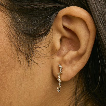 Close-up side view of a model wearing a yellow metal earring featuring alternating round white gemstones in prong settings and baguette-shaped white gemstones in bezel settings arranged in a curved line.
