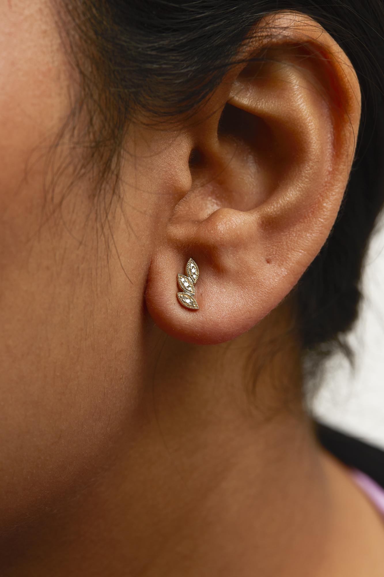 Close-up side view of a yellow metal earring on a model's ear, featuring three marquise-shaped segments each adorned with white pave-set round gemstones.