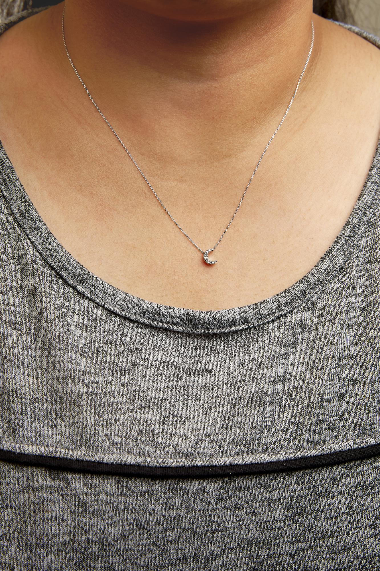 Close-up front view of a white metal necklace with a crescent moon pendant adorned with small white round pave set gemstones, worn on a person's neck against a gray knit top.