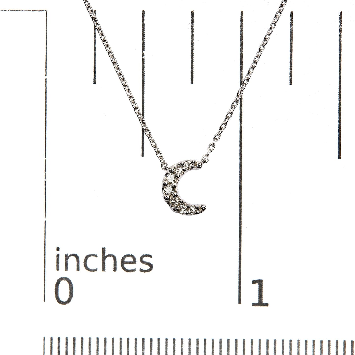 Close-up front view of a white metal necklace with a crescent moon pendant featuring multiple round white gemstones in pave settings, displayed on a ruler for size reference.