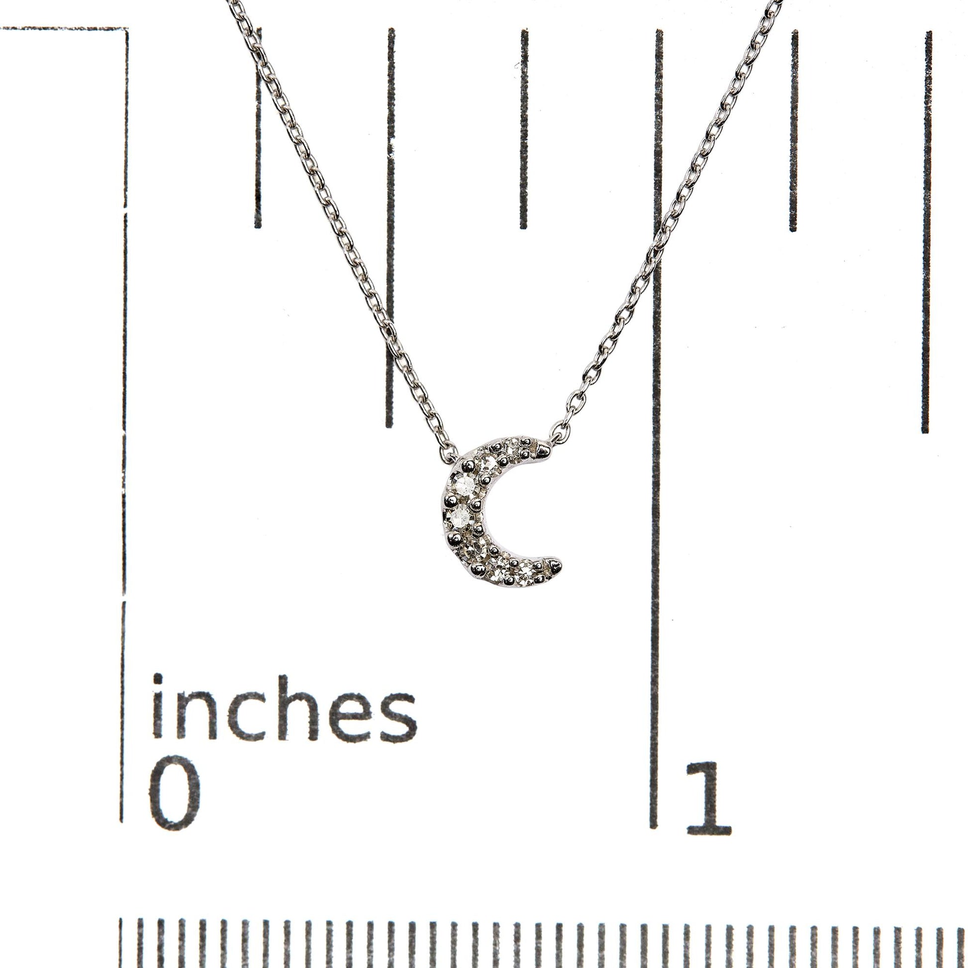 Close-up front view of a white metal necklace with a crescent moon pendant featuring multiple round white gemstones in pave settings, displayed on a ruler for size reference.