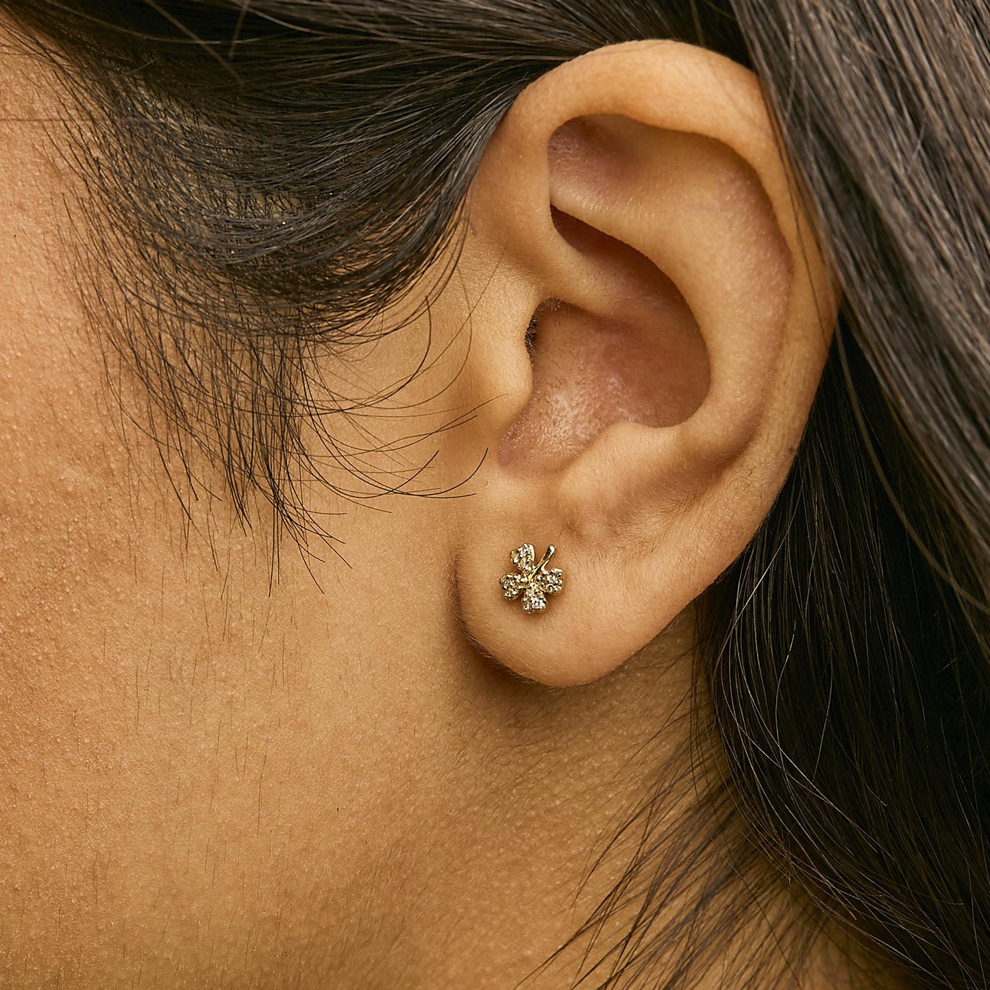 Close-up side view of a model wearing small yellow metal earrings shaped like a four-leaf clover with white gemstones in prong settings.