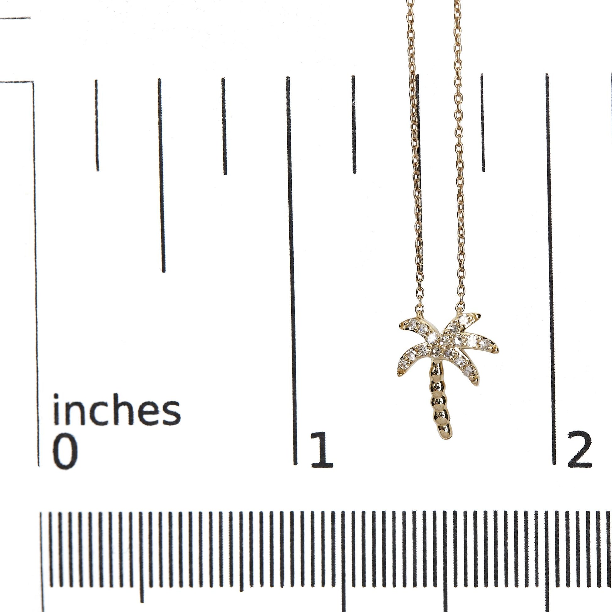 Close-up front view of a yellow metal necklace with a palm tree-shaped pendant featuring round white gemstones in pave setting, photographed on a ruler for size reference.