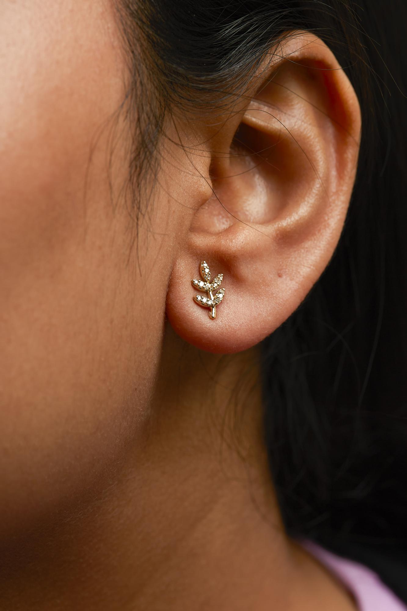 Close-up side view of a model's ear wearing yellow metal earrings shaped like a small branch with multiple leaves, each leaf set with white round pave-set gemstones.