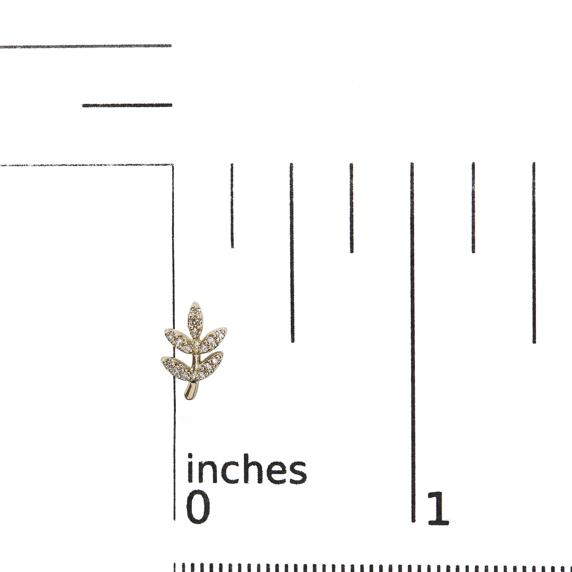 Close-up front angle of a yellow metal earring shaped like a small branch with multiple leaves paved with round white gemstones, placed on a ruler for size reference.
