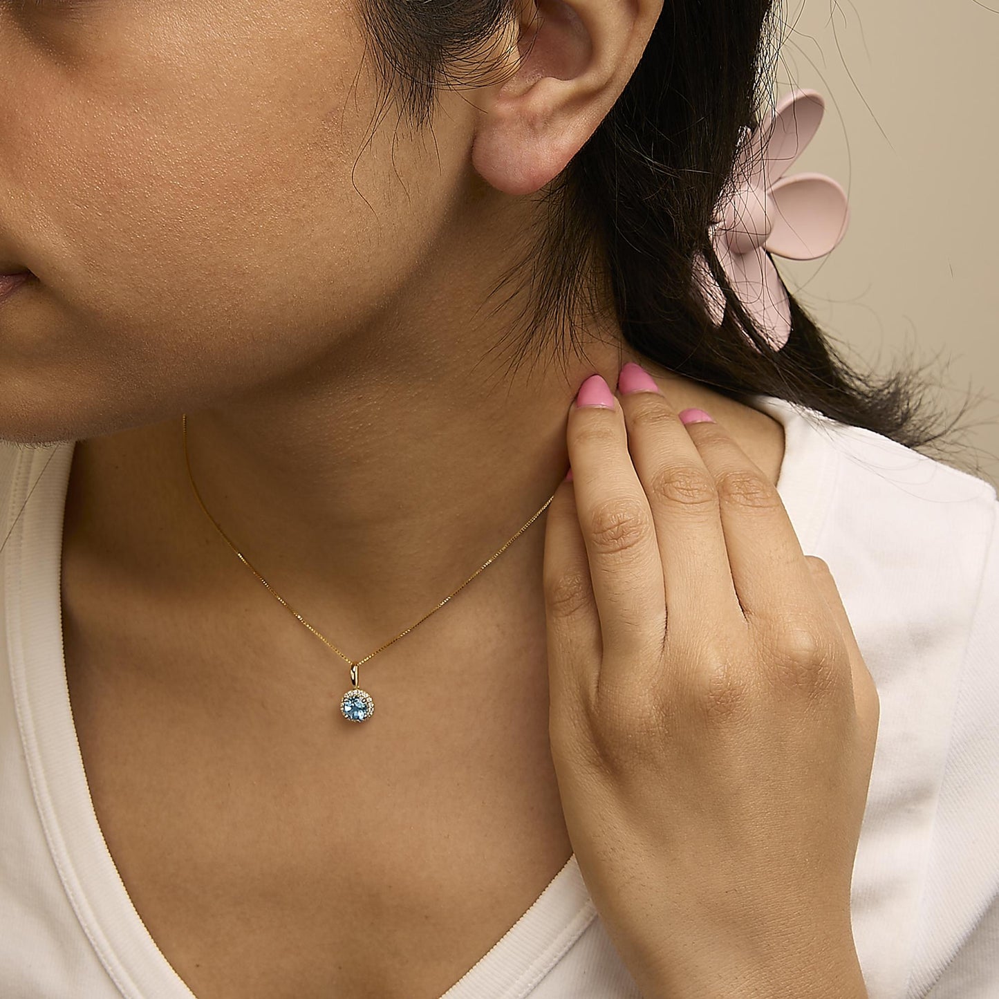 Close-up front angle of a model wearing a yellow metal necklace with a round blue gemstone pendant surrounded by small white gemstones in a shared prong setting. The model's hand is gently touching the necklace.