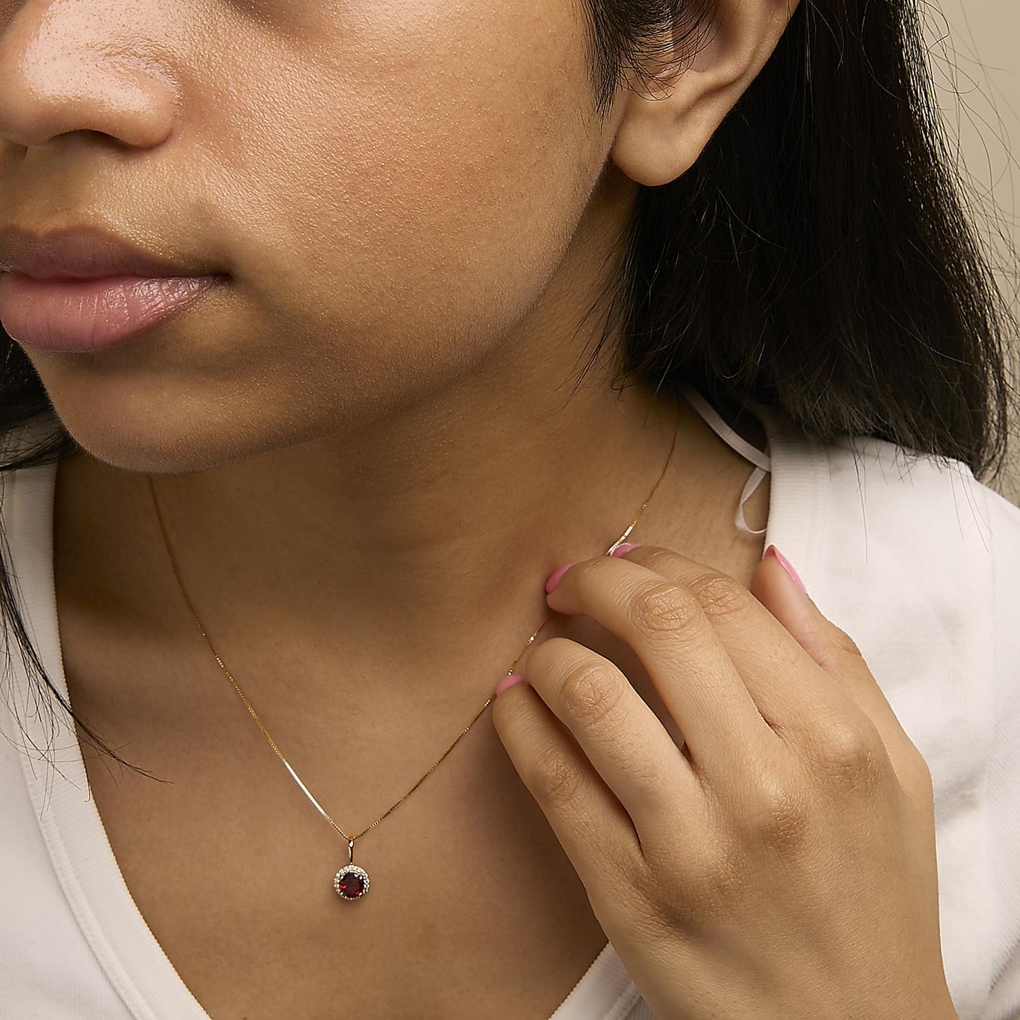 Close-up front angle of a model wearing a yellow metal necklace with a round red gemstone pendant surrounded by small white gemstones, shown with the model’s hand near the pendant.