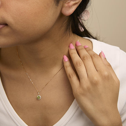 Close-up angled shot of a model wearing a yellow metal necklace with a round green gemstone pendant surrounded by small white gemstones. The model’s hand is touching the neckline area.