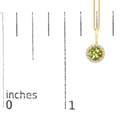 Close-up front view of a yellow metal necklace featuring a round green gemstone set in a four-prong setting, surrounded by a halo of small round white gemstones in shared prong settings, shown hanging on a scale with inch markings.