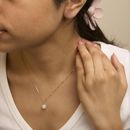 Yellow metal necklace with a round pendant featuring a central white gemstone surrounded by small white gemstones in a shared prong setting, shown worn on a model from a side angle.