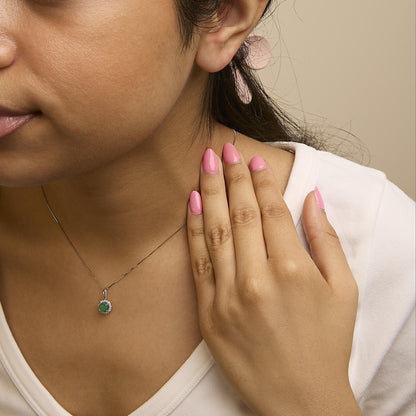 Close-up angled shot of a model wearing a white metal necklace with a round green gemstone pendant surrounded by multiple round white gemstones in a shared prong setting. The model’s hand with pink polished nails is gently touching the neck near the necklace.