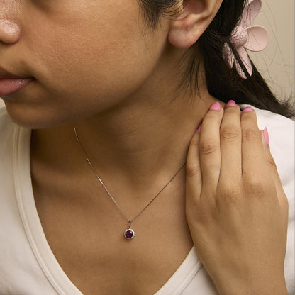 Close-up angled shot of a model wearing a white metal necklace featuring a round red gemstone pendant surrounded by small round white gemstones in a shared prong setting.