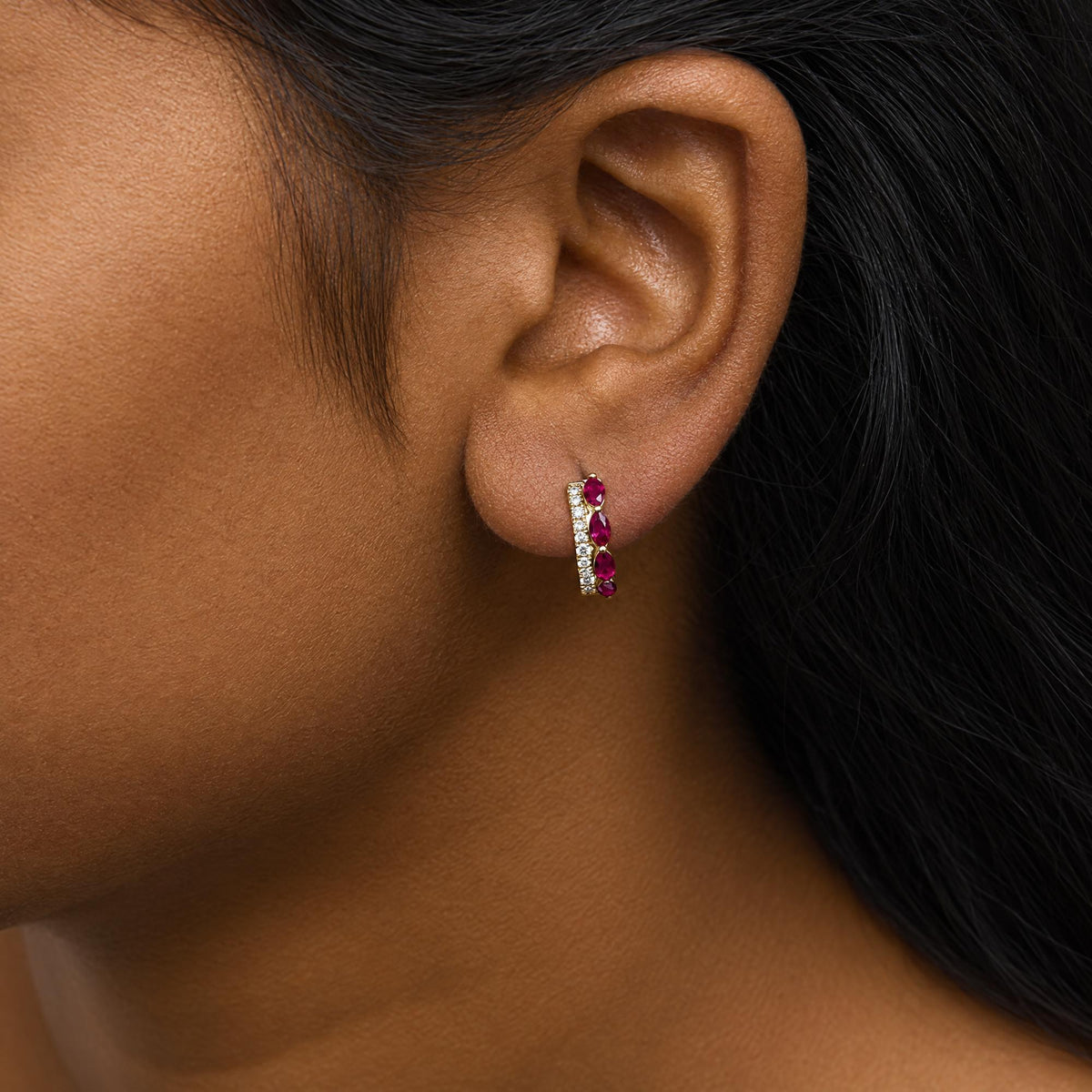 Close-up side view of a model wearing yellow metal earrings featuring marquise-shaped red gemstones and round white gemstones in prong settings.