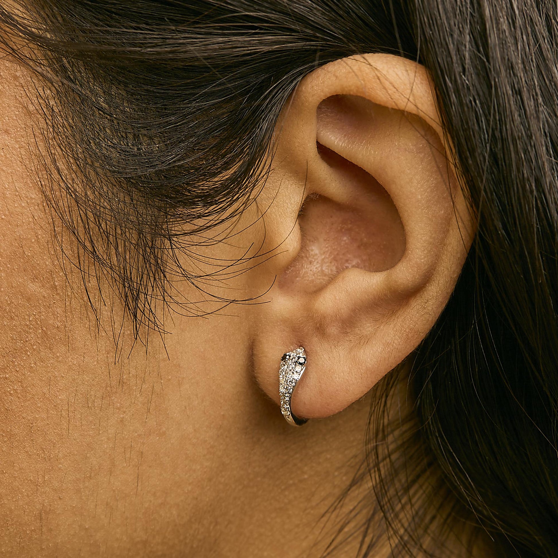 Close-up side view of a model's ear wearing a white metal earring with white and black round gemstones in a curved design.
