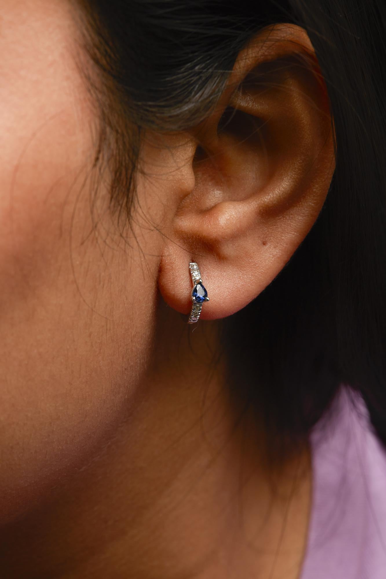 Close-up side view of white metal hoop earring on a model's ear featuring a pear-shaped blue gemstone and small round white gemstones in pave setting.