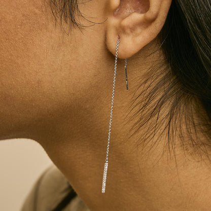 Close-up side view of a model wearing white metal dangle earrings with a thin chain and a vertical bar featuring micro-pave white gemstones.