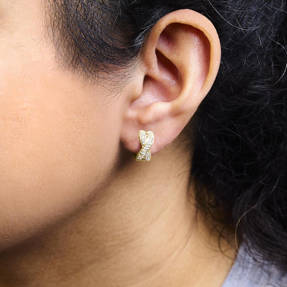 Close-up side view of a yellow metal earring on a model's ear featuring a crisscross design with round white gemstones set in a pave setting.