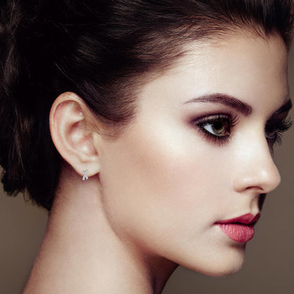 Close-up side profile of a model wearing white metal earrings with an X shape, featuring round white gemstones in prong settings.