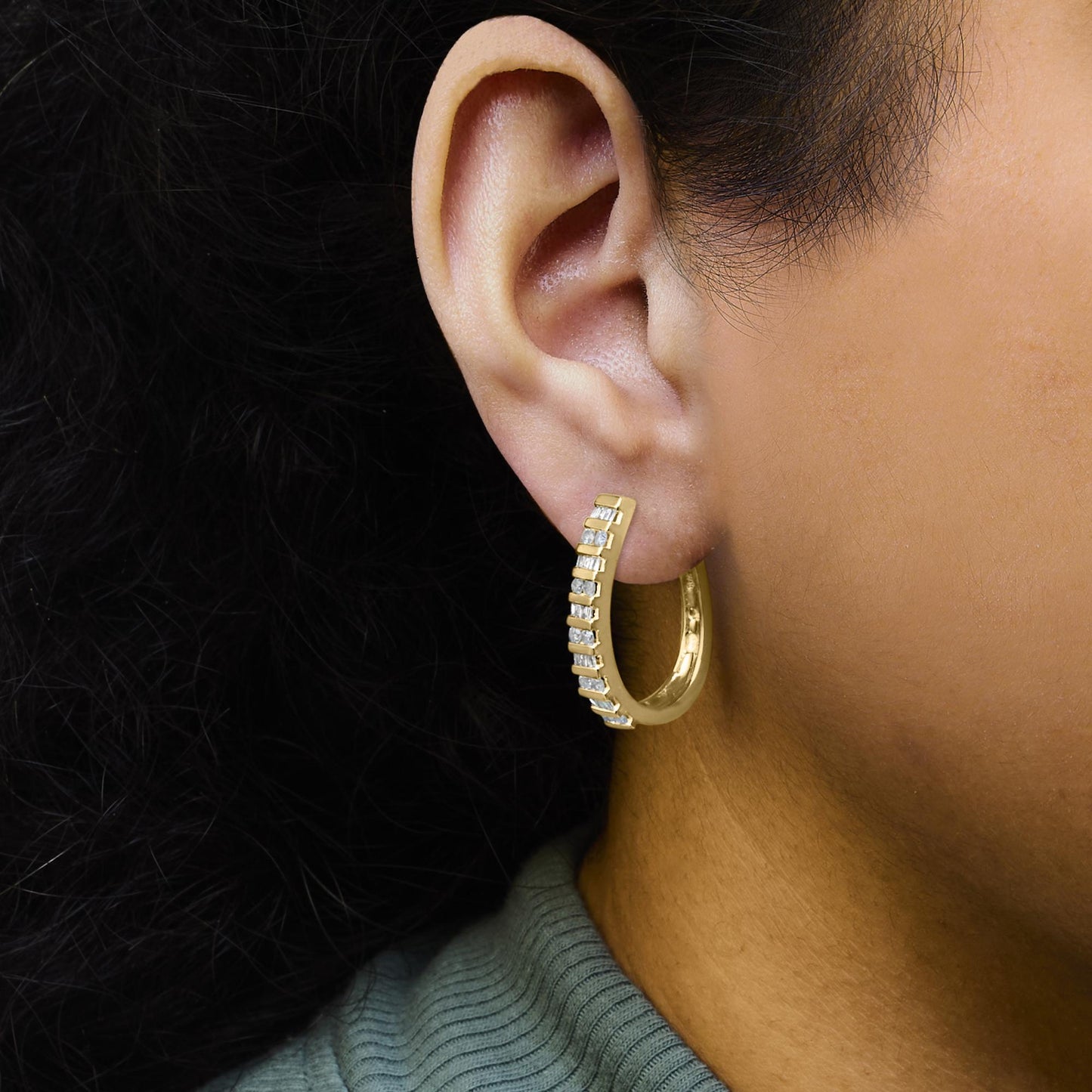 Close-up side view of a yellow metal hoop earring on a model's ear, featuring channel-set white round and baguette-shaped gemstones along the front curve.