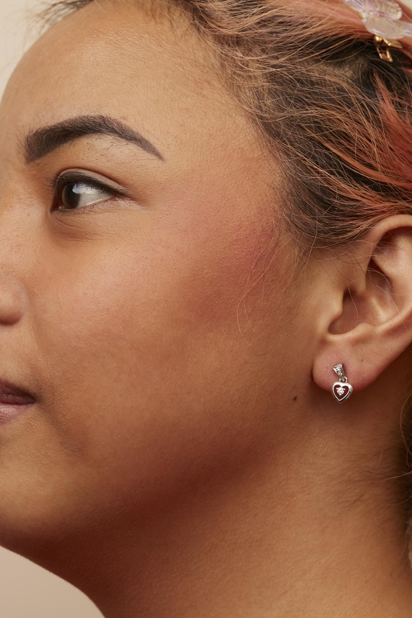Close-up side view of a model wearing white metal heart-shaped earrings with prong-set round white gemstones.