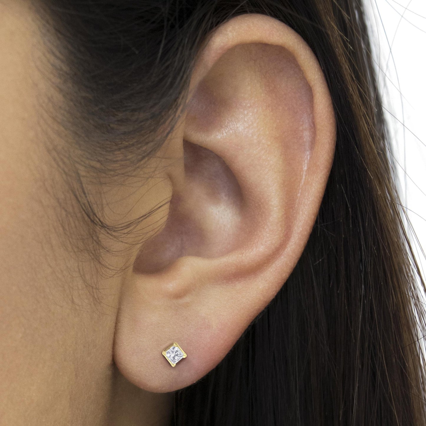 Close-up side view of a yellow metal earring with a princess-shaped white gemstone in a four-prong setting, worn on a model's ear.