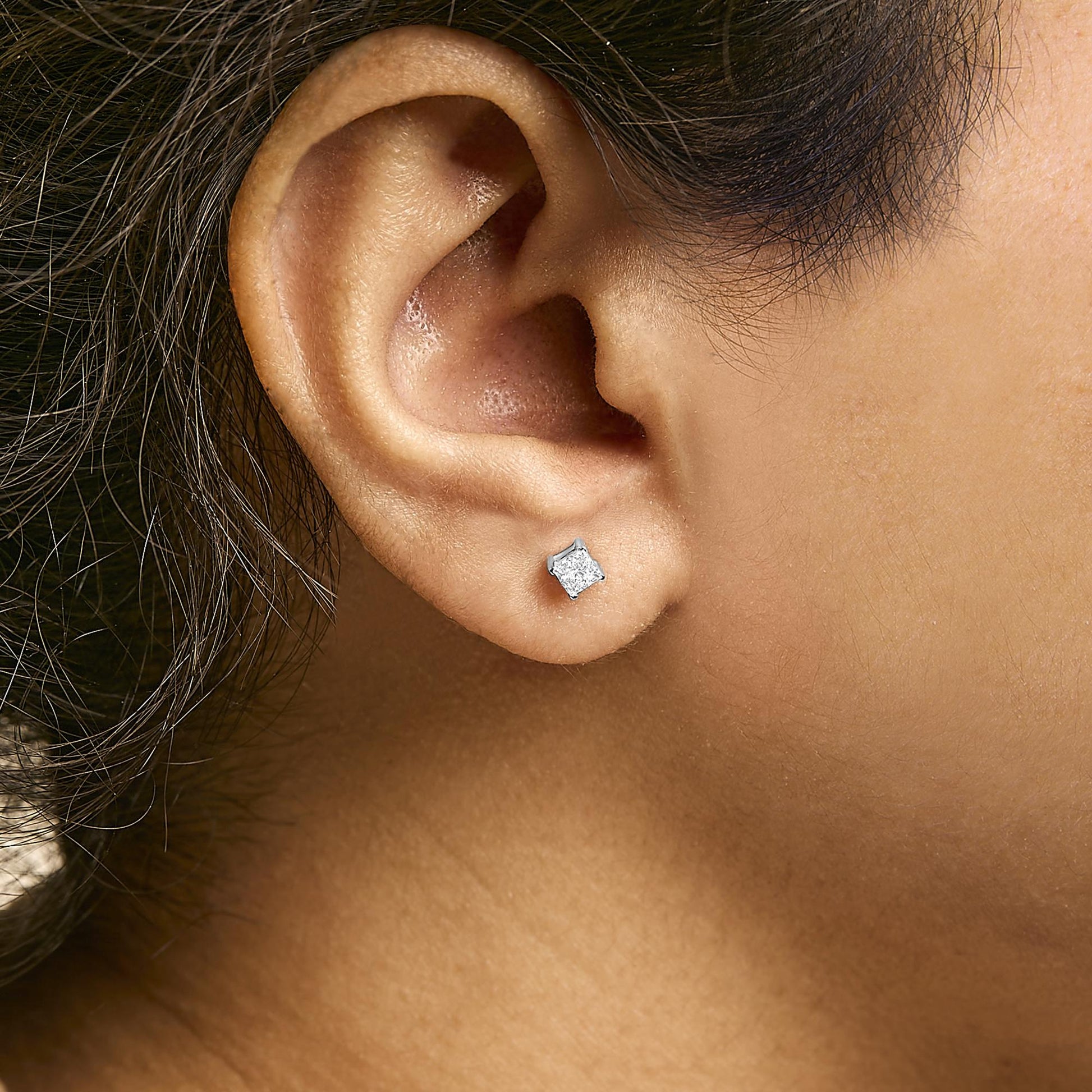 Close-up side view of a white metal earring with a princess-shaped white gemstone in a four-prong setting, worn on a model's ear.