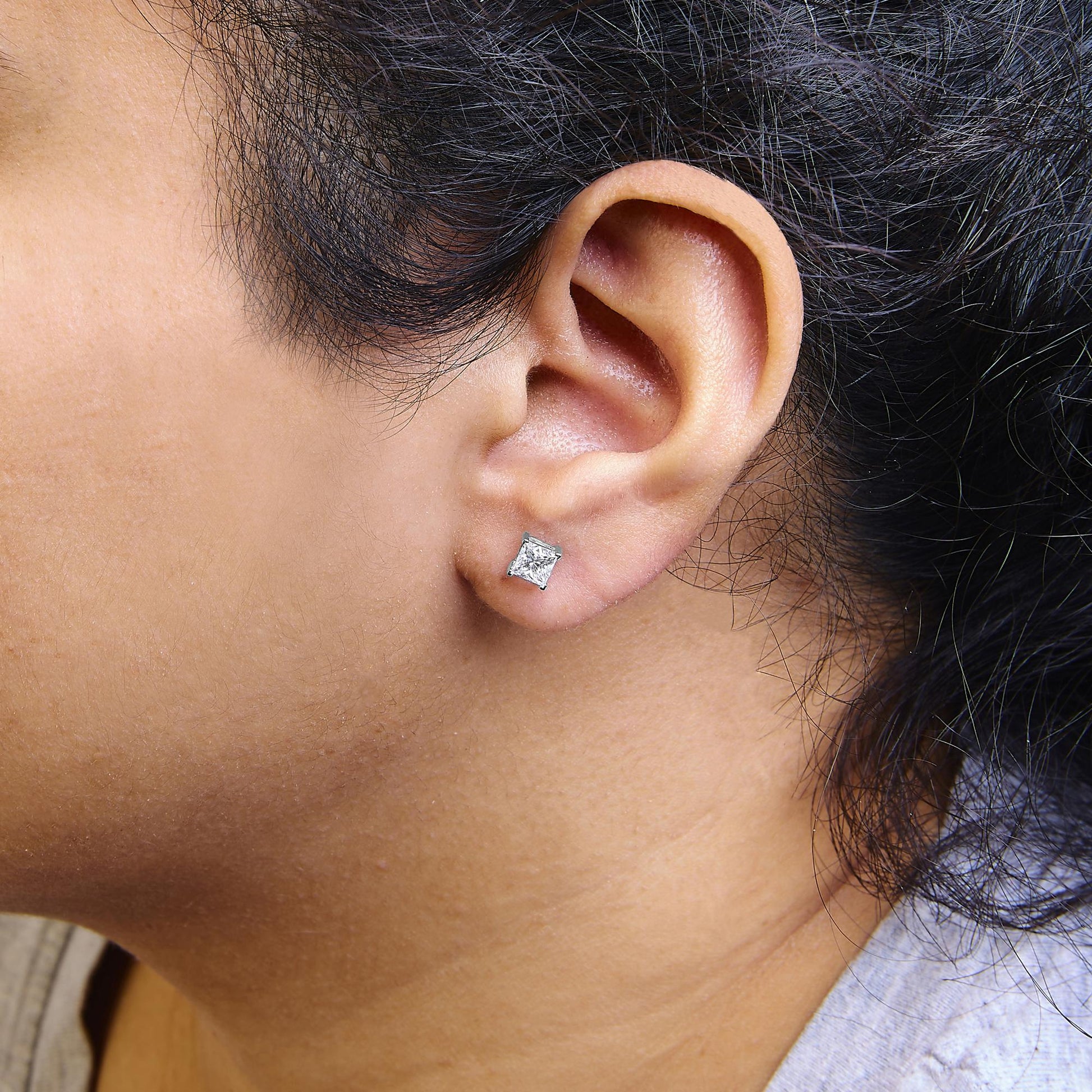 Close-up side view of a white metal stud earring with a princess-shaped white gemstone in a four-prong setting worn on a model's ear.