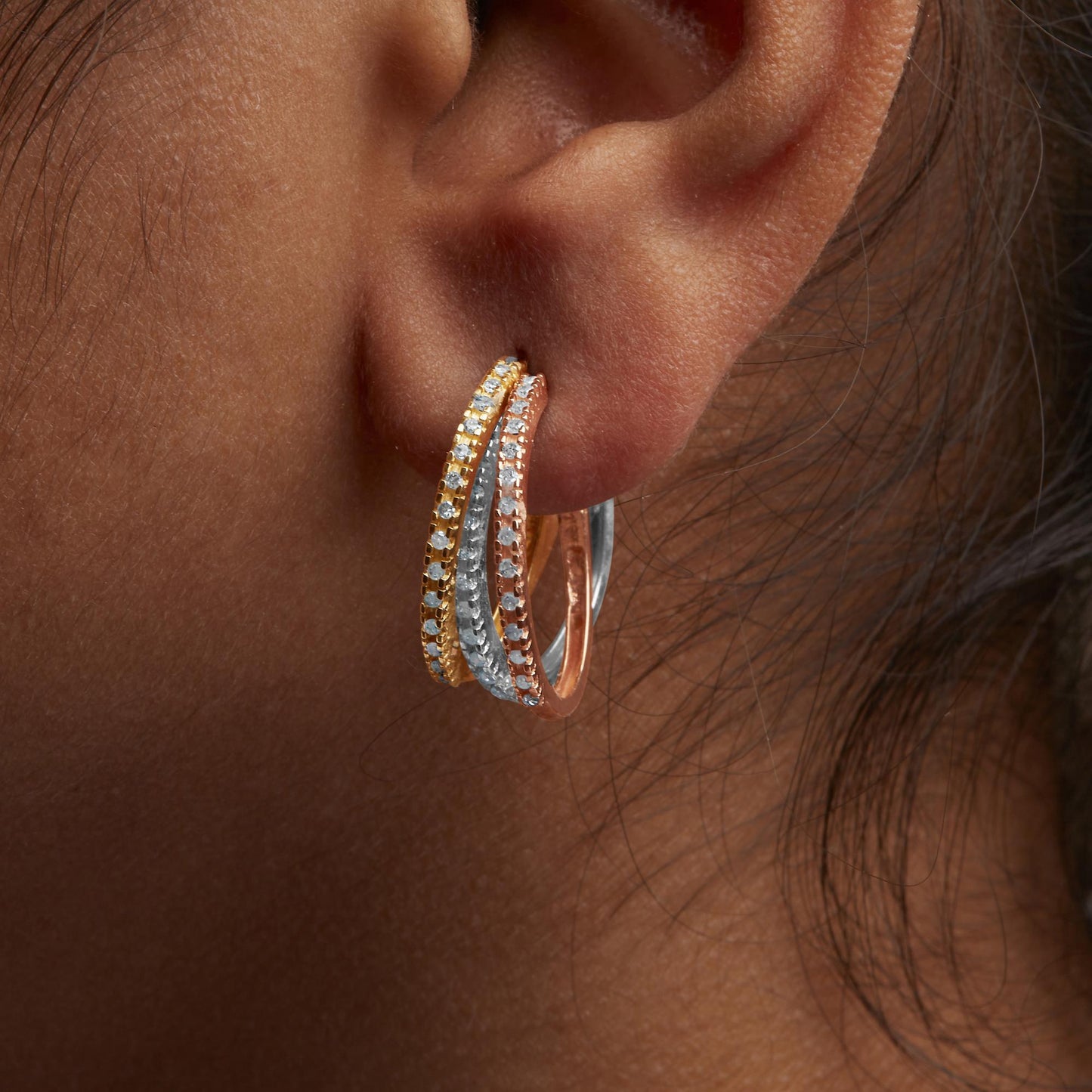 Close-up side view of a person's ear wearing a tri-color hoop earring with yellow, white, and rose metal bands, each set with round white gemstones in prong settings.