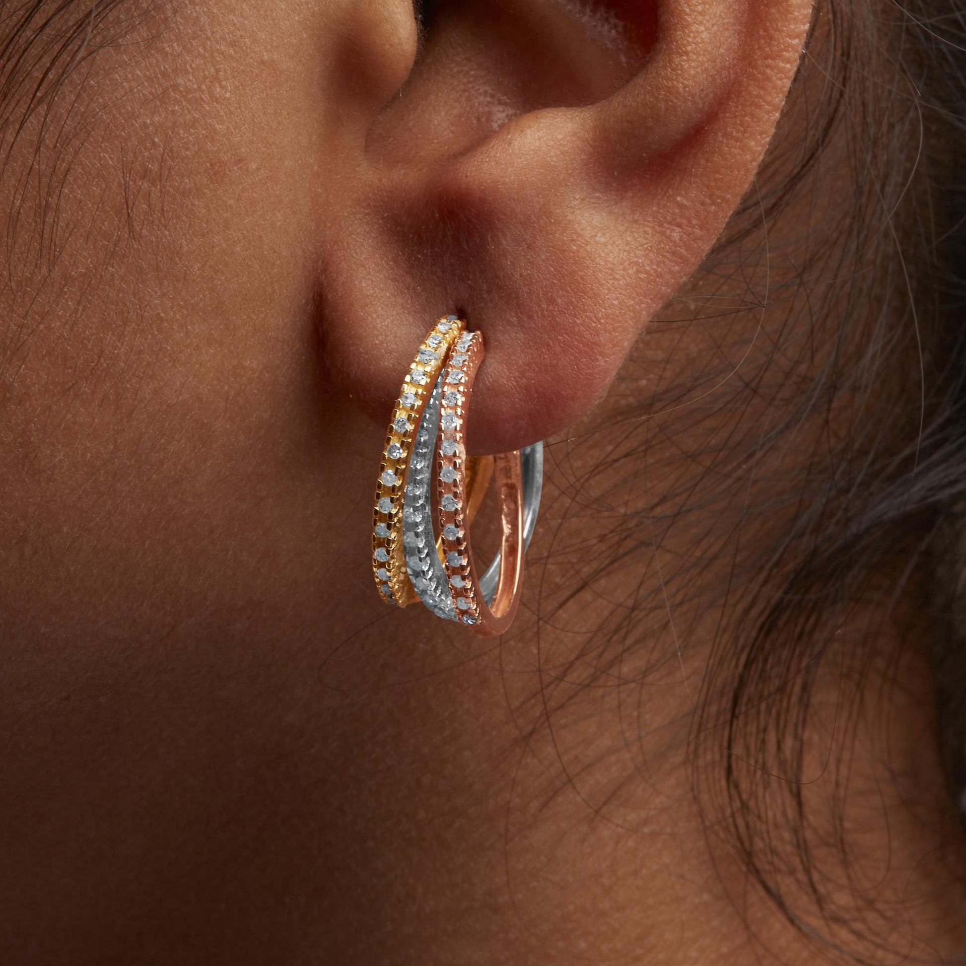 Close-up side view of a person's ear wearing a tri-color hoop earring with yellow, white, and rose metal bands, each set with round white gemstones in prong settings.