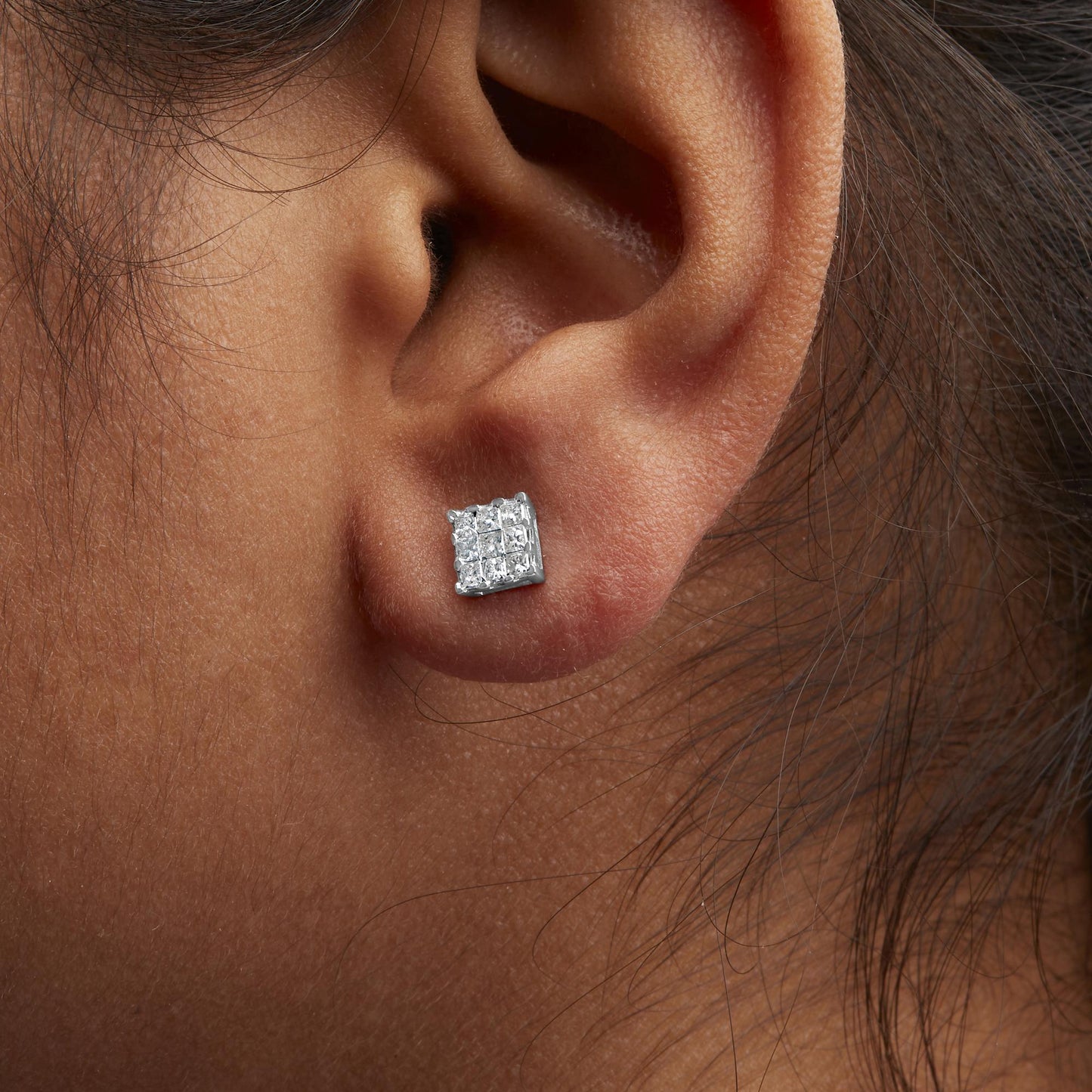 Close-up side view of a white metal stud earring with a cluster of princess shape white gemstones in an invisible setting, worn on a model's ear.