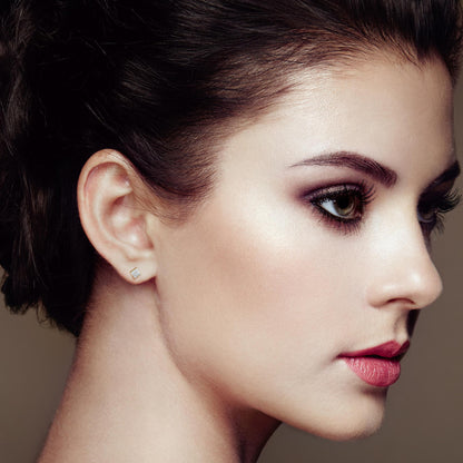 Close-up side profile of a model wearing small square-shaped yellow metal stud earrings with invisible-set white baguette and princess-shaped gemstones.