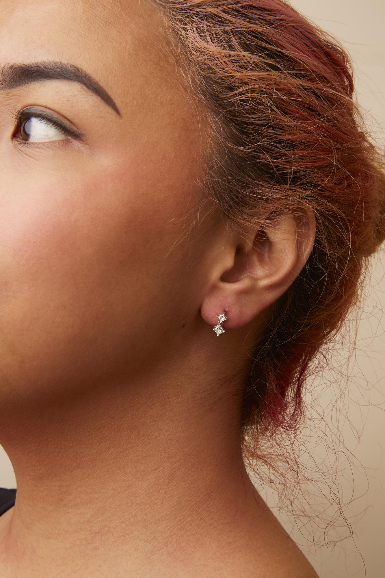 Close-up side view of a model wearing white metal earrings featuring two princess shape white gemstones in prong settings.
