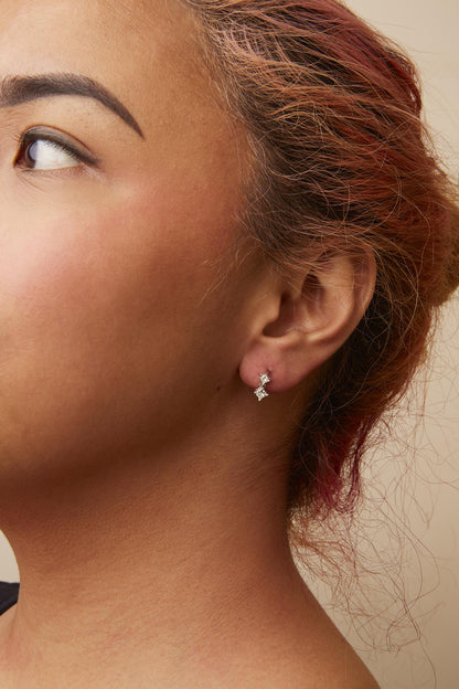 Close-up side view of a model wearing white metal earrings featuring two princess shape white gemstones in prong settings.
