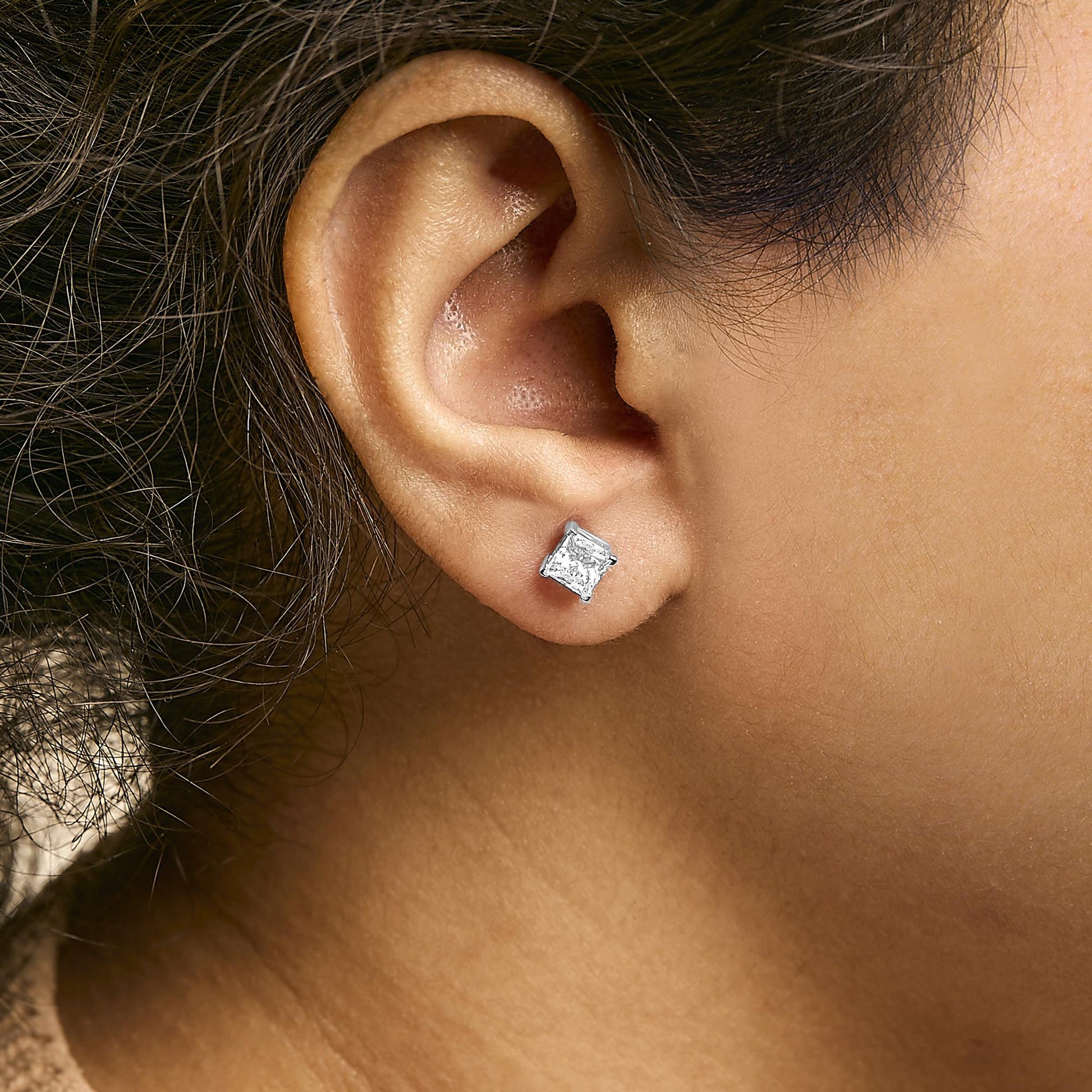 Close-up side view of a white metal earring with a princess-shaped white gemstone in a 4-prong setting worn on a model's ear.