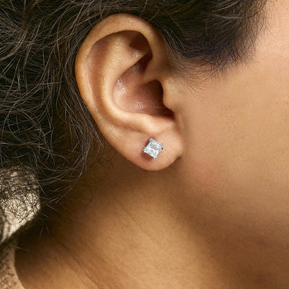 Close-up side view of a white metal earring with a princess-shaped white gemstone in a 4-prong setting worn on a model's ear.