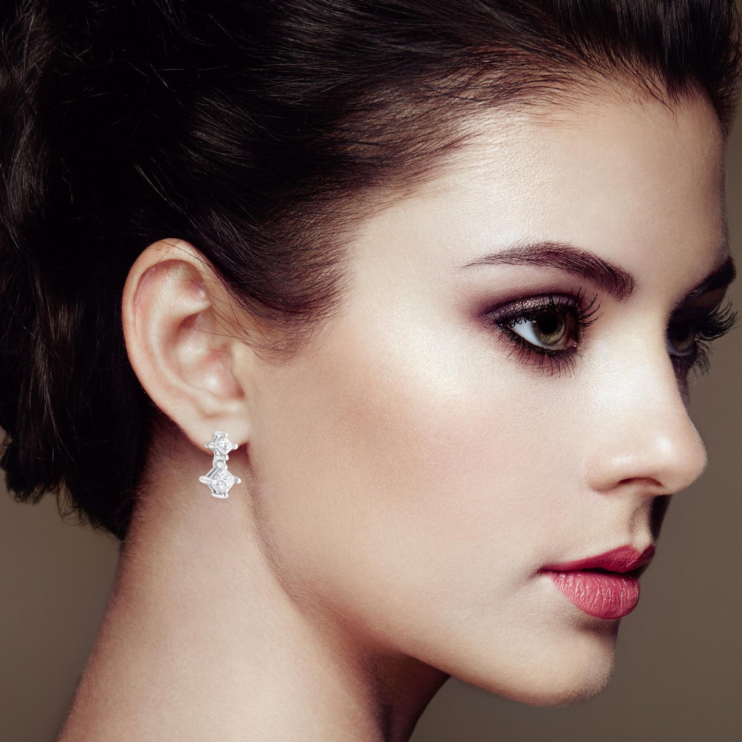 Close-up side profile of a woman wearing white metal earrings featuring two prong-set, princess-shaped white gemstones on each earring.