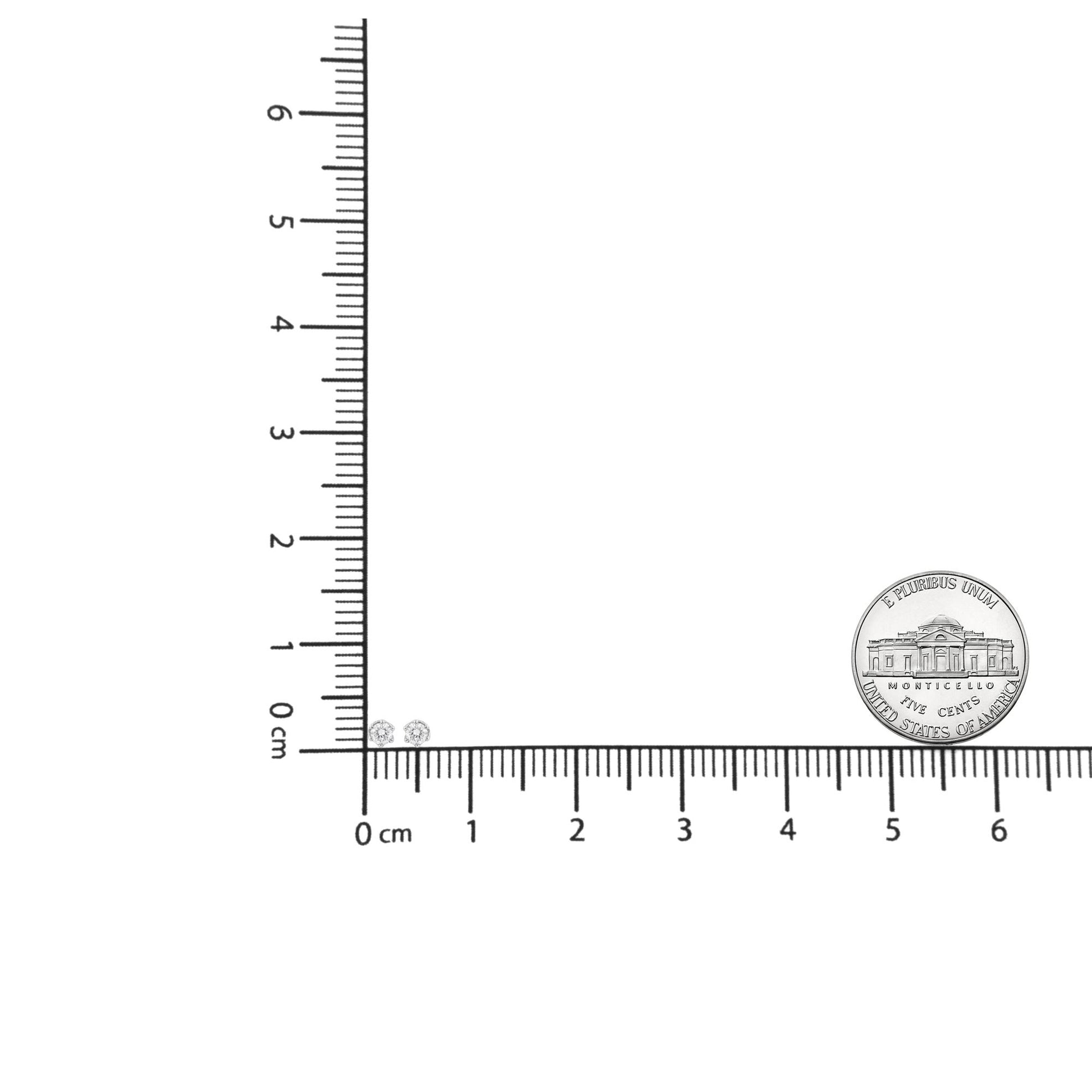 Pair of white metal round stud earrings with central round gemstone in 6-prong setting, surrounded by pave-set smaller round white gemstones, shown from front angle on ruler for size reference with coin nearby.